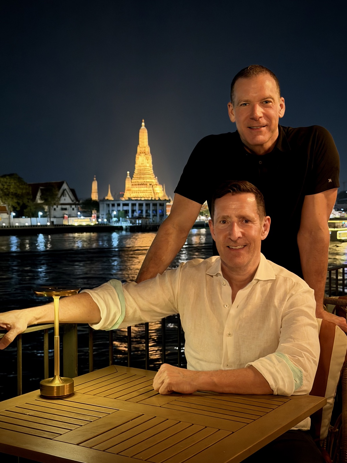 Greg and Shawn at a riverside table in Bangkok, Wat Arun glowing behind them